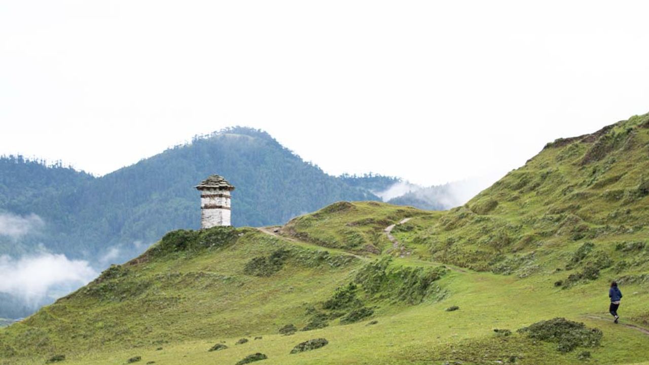 Rolling green hills of Phobjikha Valley with a traditional Bhutanese watchtower, misty mountains in the background, and a lone traveler walking along a quiet trail