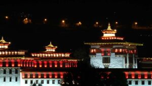 Majestic Bhutan monastery illuminated at night, with traditional white walls and red-lit windows glowing against a dark hillside backdrop.