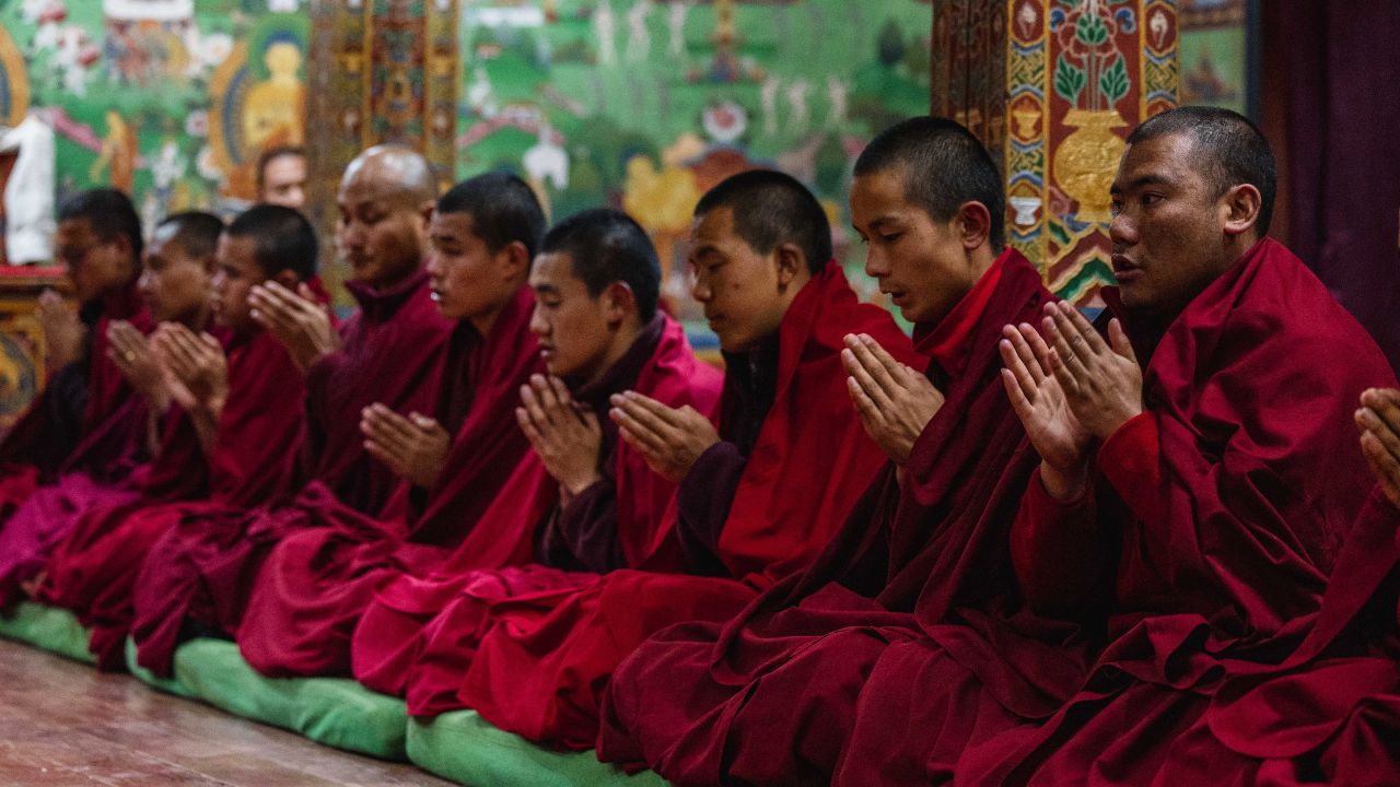 Buddhist monks in maroon robes seated in a row inside a richly decorated monastery, praying with folded hands before colorful wall murals and ornate pillars.