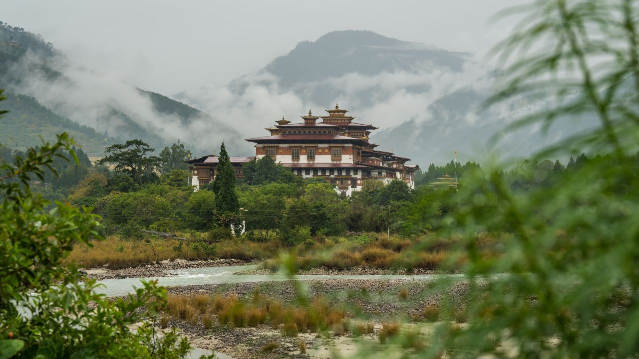 Bhutanese monastery surrounded by greenery, river, and misty mountains.