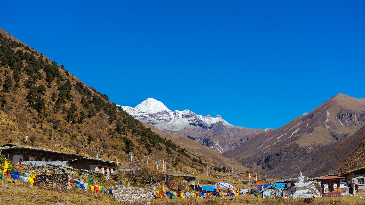 Snow-capped Himalayan peak rising above a remote mountain village with colorful prayer flags, stone buildings, and brown alpine slopes under a clear blue sky.