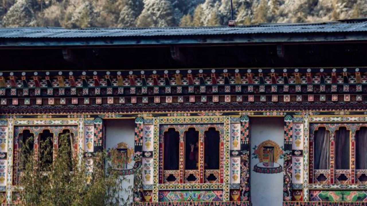 Traditional Bhutanese building with colorful painted windows set against snow-covered forested mountains.