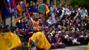 Masked dancer performing a traditional Bhutanese festival dance with drum while a crowd watches.