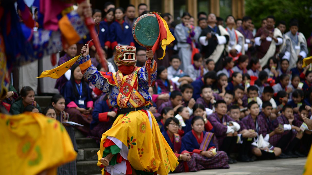 Masked dancer performing a traditional Bhutanese festival dance with drum while a crowd watches.
