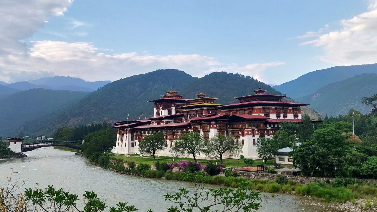 Punakha Dzong standing beside the river in Bhutan, surrounded by green hills and blooming flowers under a clear blue sky.