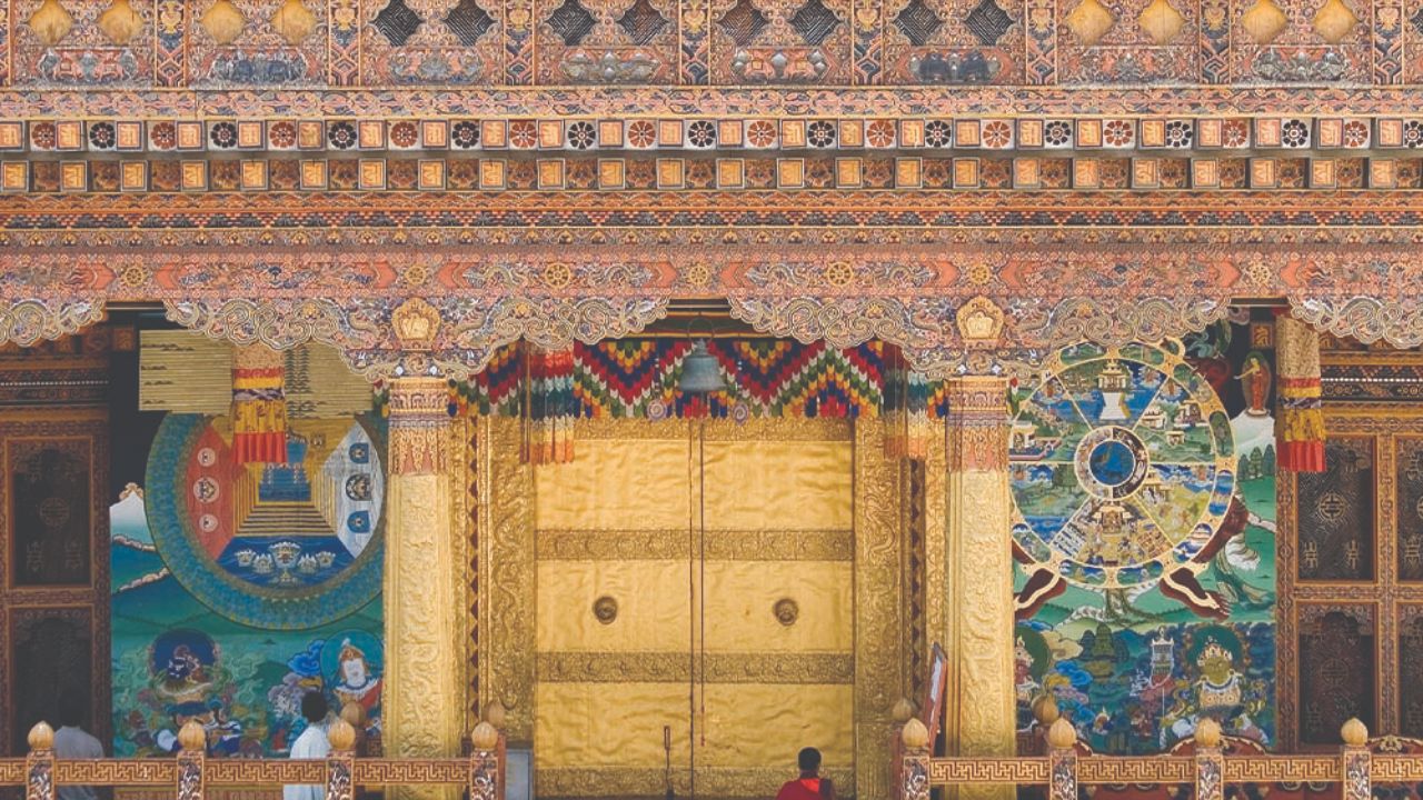 Monk in red robes before golden doors inside an ornate Bhutanese monastery.