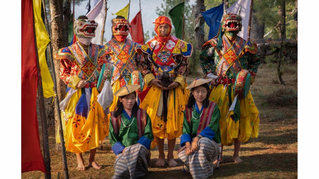 Group of Bhutanese festival performers in colorful traditional costumes and masks standing outdoors with prayer flags in the background.